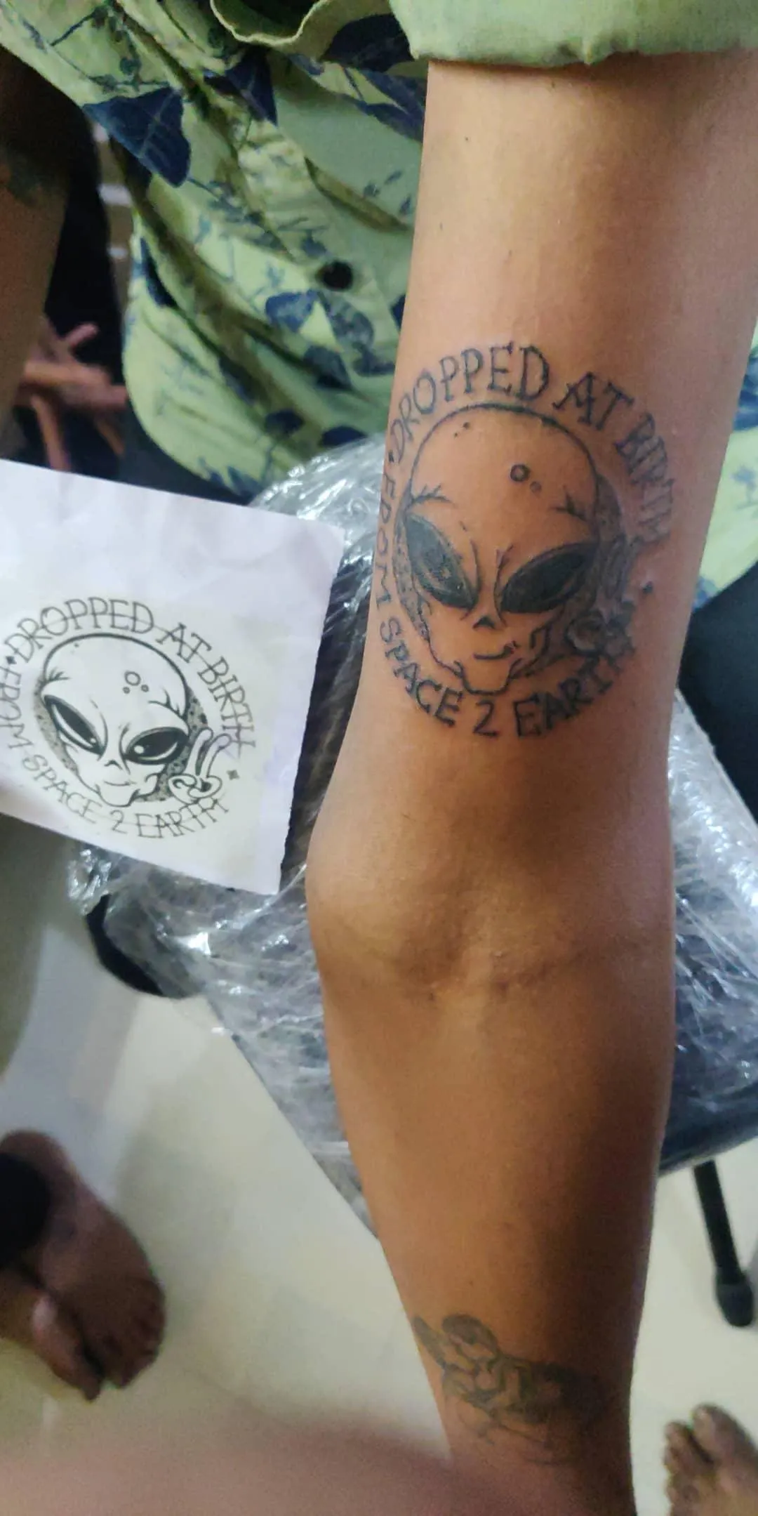 Alien-face tattoo with circular lettering on a forearm beside a printed reference design