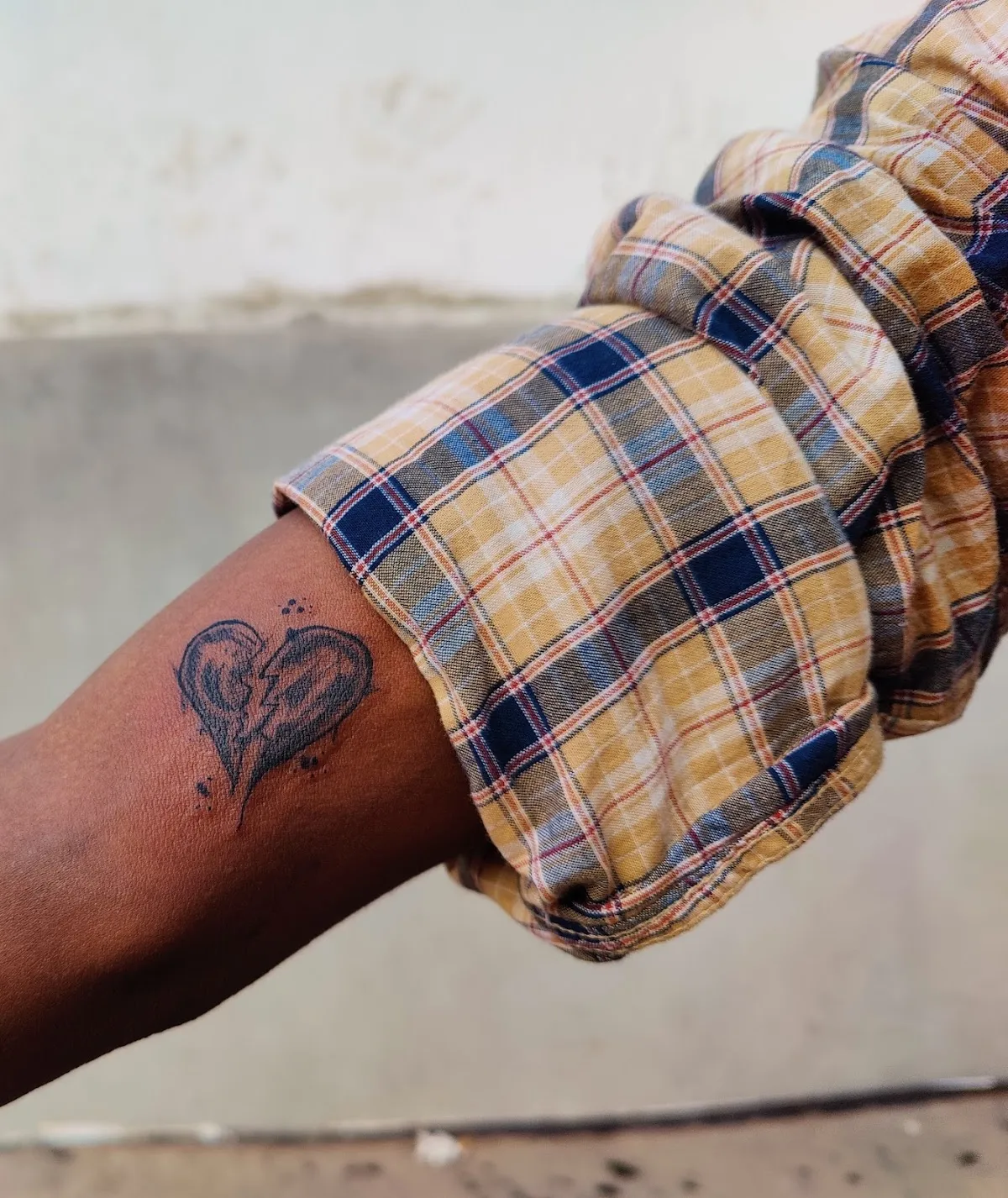 Black broken-heart tattoo on a forearm beside a rolled checked shirt sleeve