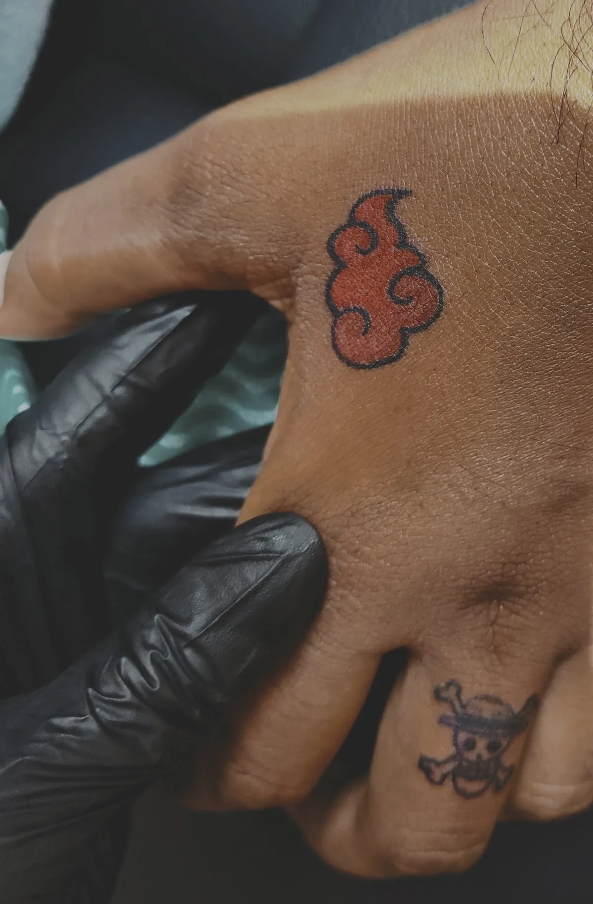 Small red cloud tattoo outlined in black on the back of a hand held by black-gloved fingers