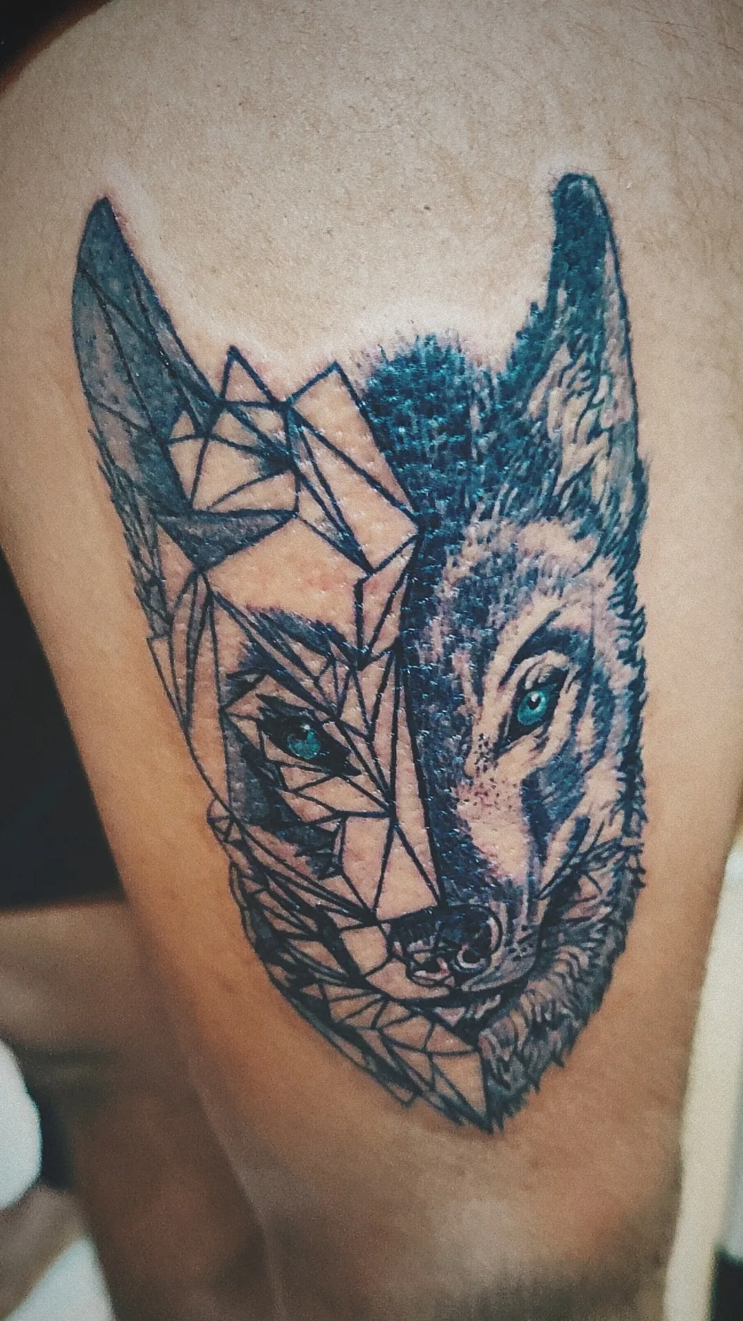 Wolf-face tattoo on skin combining angular geometric linework with shaded fur and blue-green eyes